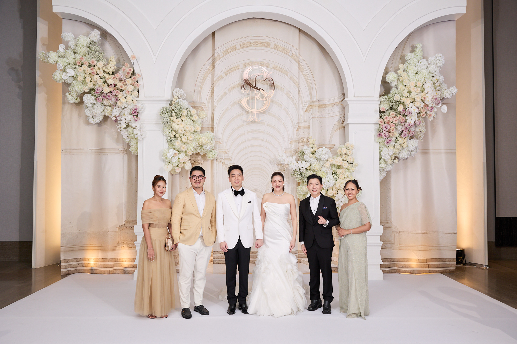 3355321 คู่บ่าวสาวและผู้ร่วมงานยืนถ่ายภาพหมู่หน้าฉากซุ้มโค้งและดอกไม้สีขาวในงานมงคลสมรส